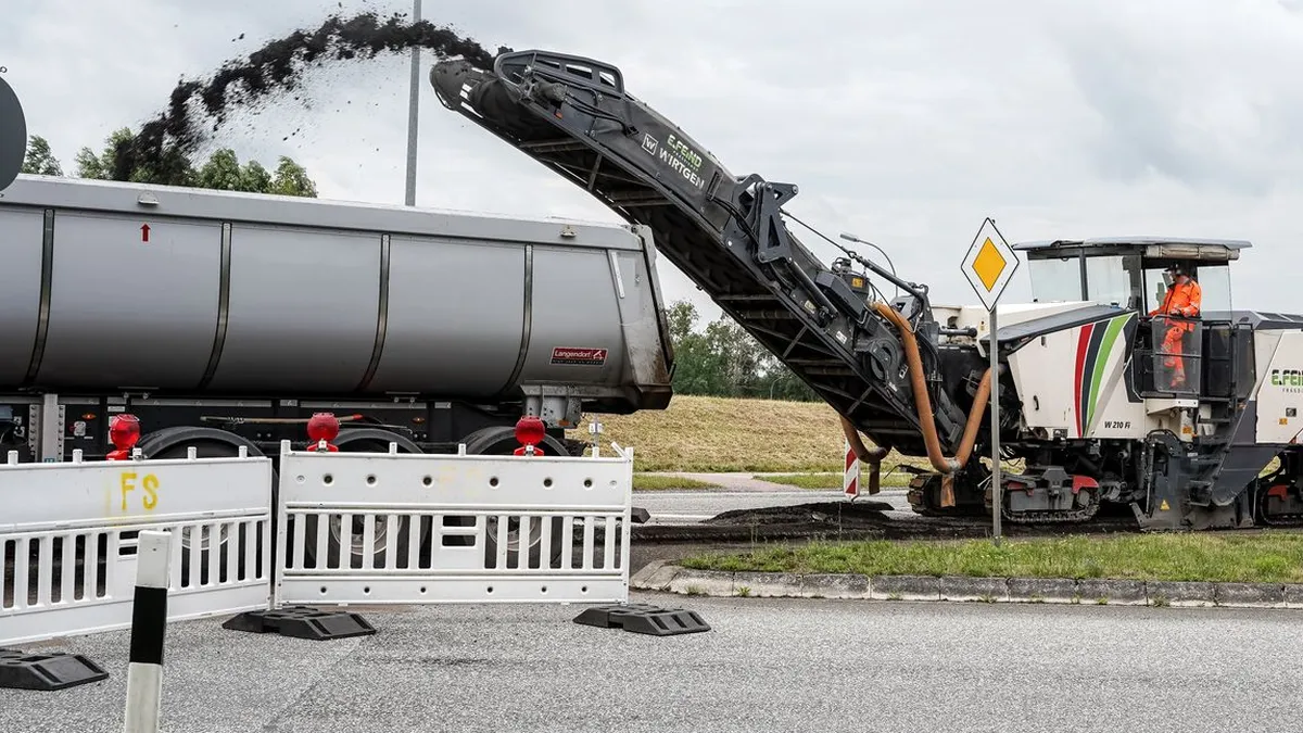 A cold milling machine grinding off the old asphalt surface and conveying the millings into a dump truck behind a work zone barrier