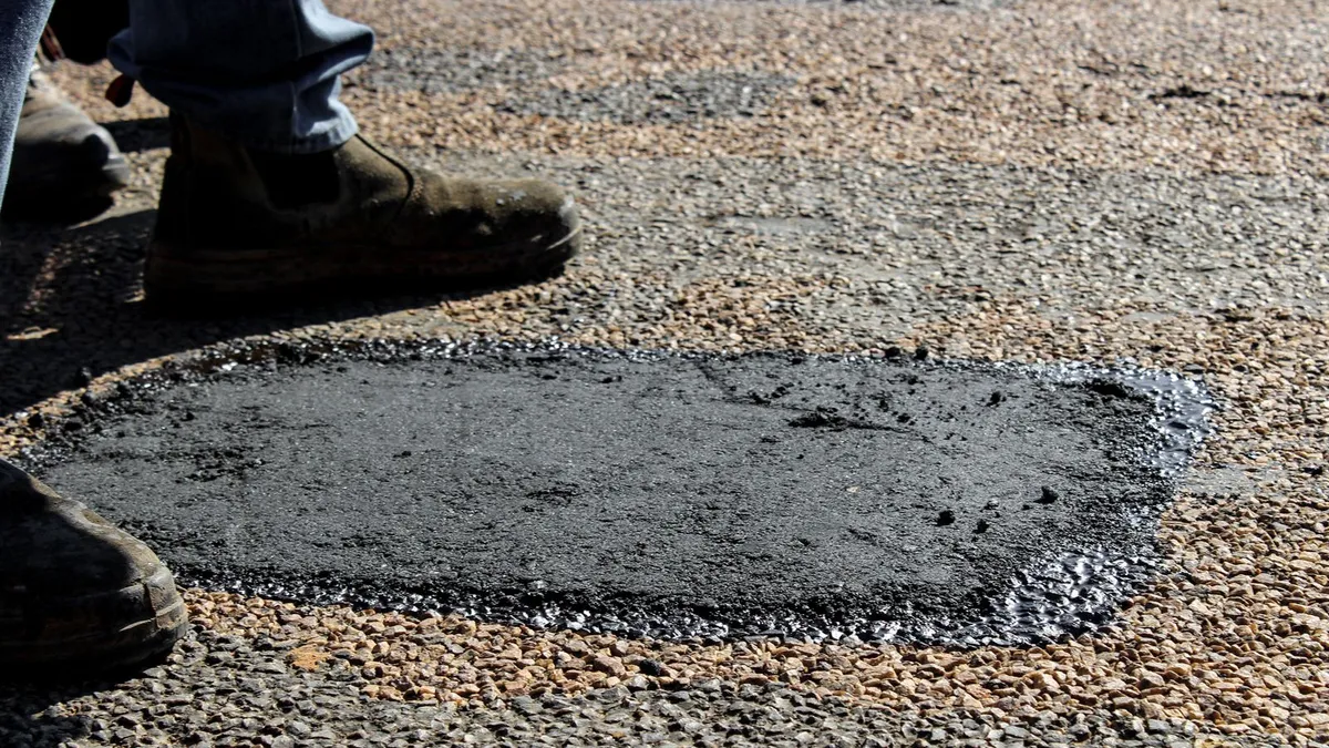 A freshly placed hot-mix patch filling a pothole in worn asphalt, with a worker boots standing at the edge of the repair