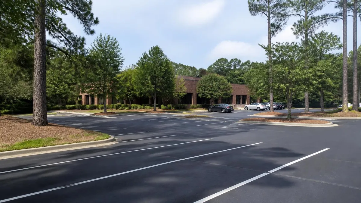 A freshly sealed and striped office park parking lot in suburban Atlanta, shaded by tall loblolly pines, with a low brick office building behind