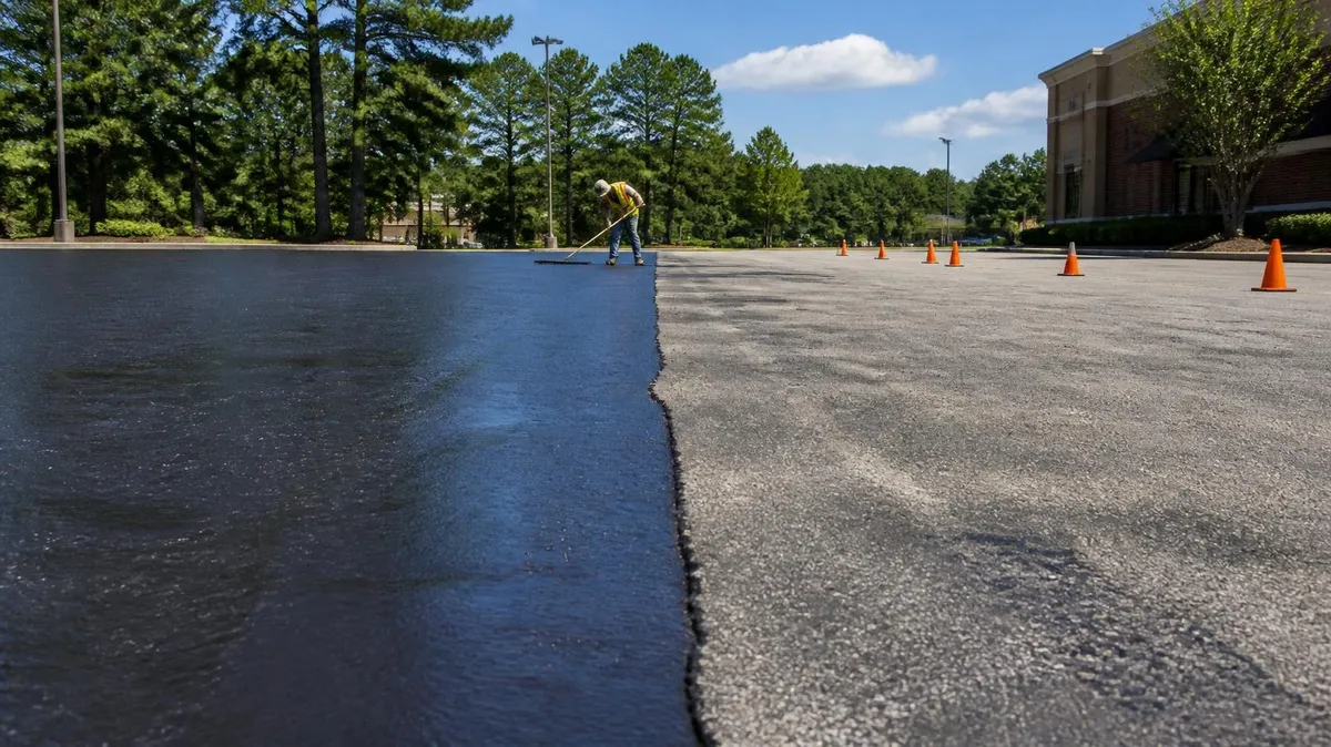 A commercial parking lot mid-sealcoat, fresh glossy black sealer meeting faded gray asphalt along a straight edge, a worker with a squeegee and orange cones beyond